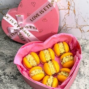 A pink heart-shaped box with a bow and "LOVE IS ETERNAL" contains eight Mango & Passion Fruit macarons with creamy filling. Lined with pink paper, it rests on a marble surface—a delightful treat evoking charming UK patisseries.