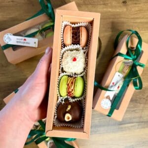 A hand holds an open cardboard box containing four decorated desserts: a macaron, a coconut-covered treat, a green dessert, and a chocolate treat with a gold garnish. The box is placed over a wooden surface with similar boxes nearby.
