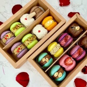 The Macarons Custom Box, showcasing an assortment of vibrant macarons with chocolate, fruit, and nut fillings, is elegantly presented on a marble surface adorned with delicate rose petals for added charm.