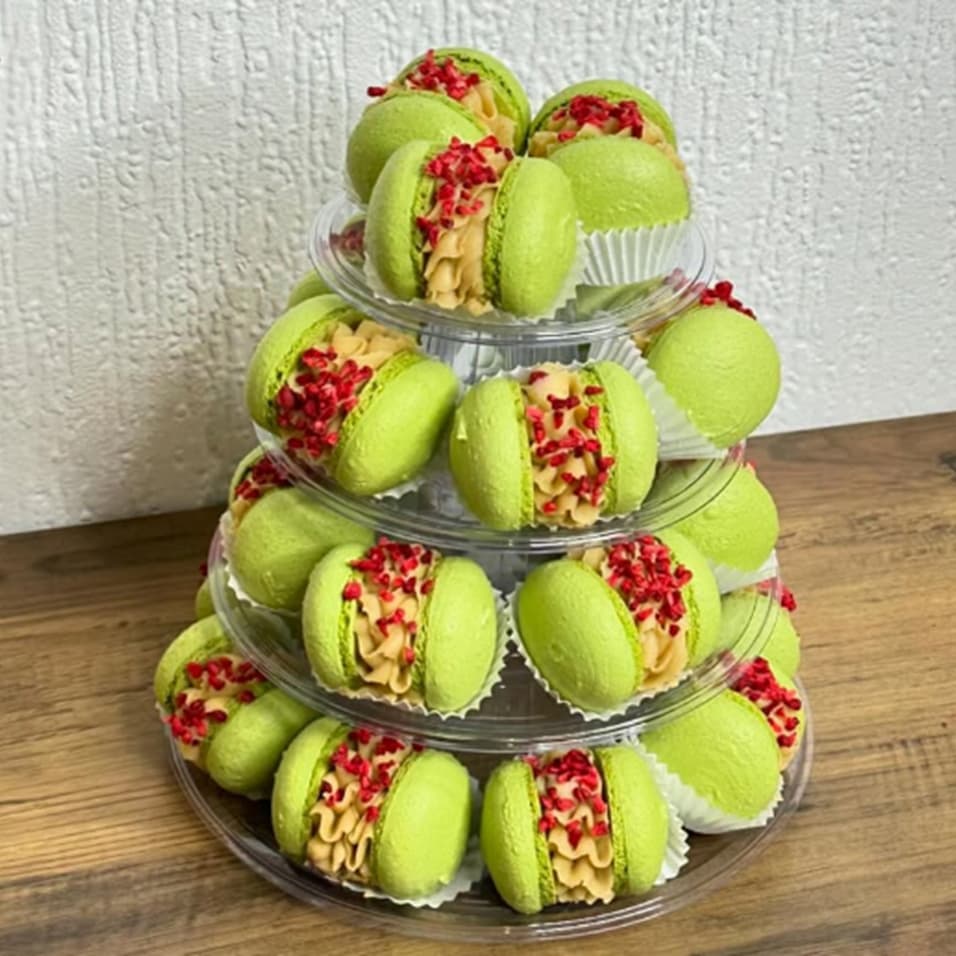 Tiered display of Pistachio Raspberry Macarons arranged semicircularly on a plastic stand against a textured white wall, featuring green macarons with cream filling and red sprinkles, creating a charming Scottish pattern on a wooden surface.