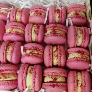 A close-up image showcases Mango Ice Cream Macarons arranged enticingly in a box. Each features a white cream filling, elegantly sprinkled with red powder. White decorative paper shreds charm from beneath. Ideal sweet indulgence for UK buyers seeking delightful treats.