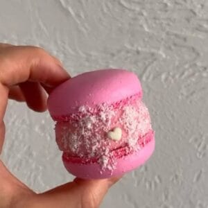 A hand holds a Raspberry Coconut Macaron with textured filling, shredded coconut, and a heart-shaped decoration against a plain white wall, inviting you to indulge in the finest macarons available for purchase in the UK.
