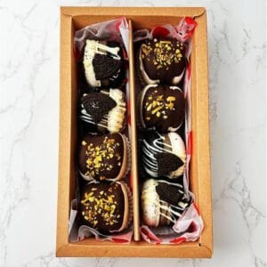 A box of Black-White Macarons with the new OREO flavor is elegantly arranged in two rows. The sleek brown paper box rests on a white marble surface.
