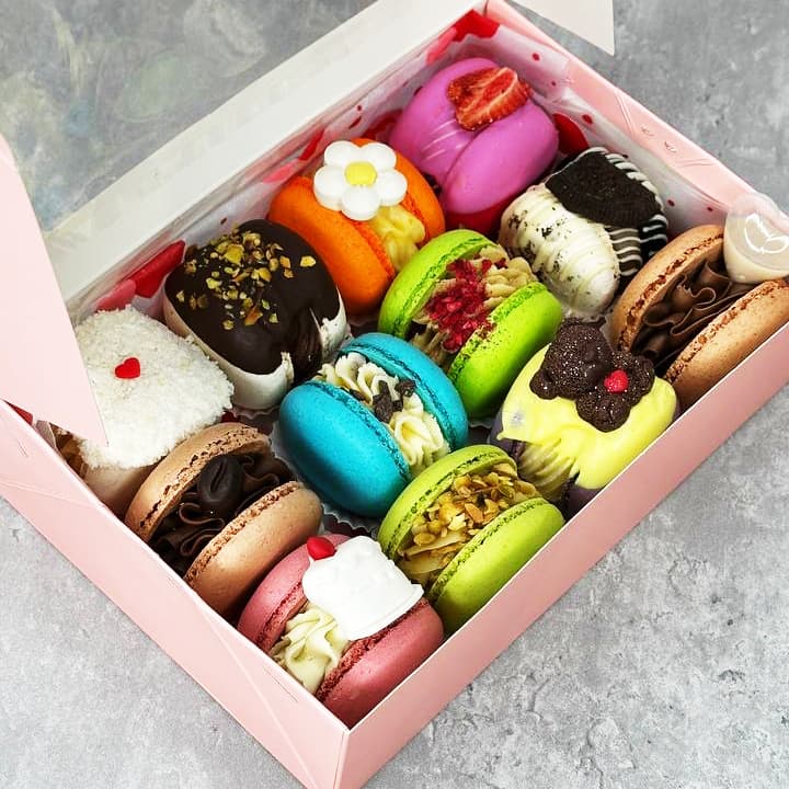 A pink box containing assorted decorated macarons with various toppings such as chocolate, flowers, berries, and nuts, arranged in rows. The colors include pink, green, blue, and orange, placed on a gray marble surface.