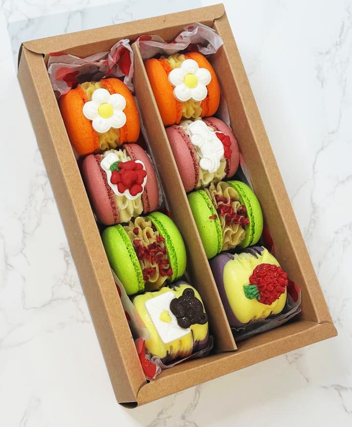 A box of macarons displayed on a marble surface. The macarons are in various colors, decorated with floral and fruit designs. There are also two square cakes with fruit toppings included in the box.