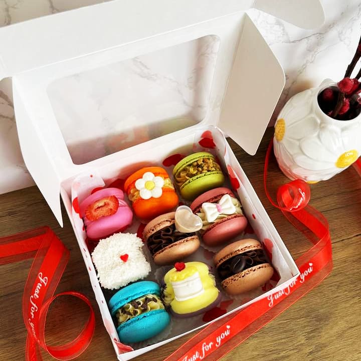 A box of nine colorful macarons, each with unique decorations, sits open on a wooden surface. A red ribbon surrounds the box. A white vase with cherry blossom branches is nearby. The macarons and decor appear playful and vibrant.