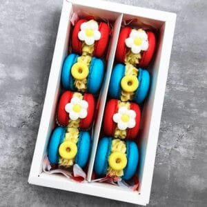 The Macarons Lacuree Box features 12 macarons in two alternating rows of red and blue, each beautifully adorned with yellow filling and topped with small white flower-shaped accents, elegantly placed on a gray surface.
