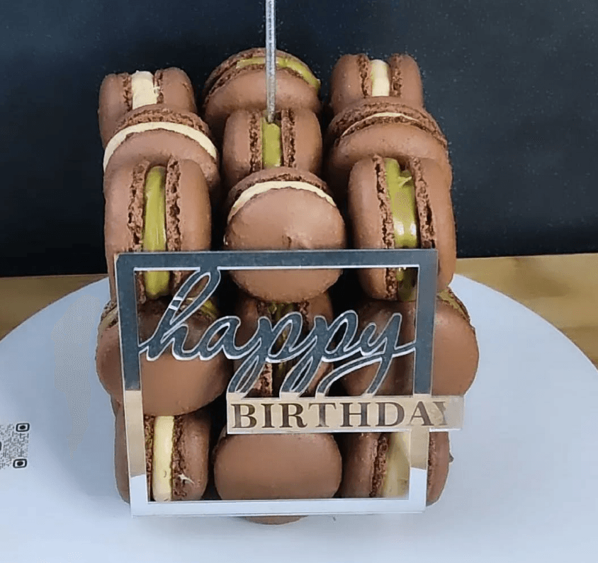 A macaron tower with layers of chocolate and green macarons is topped with a rectangular sign that reads "happy BIRTHDAY" in metallic script. The tower is placed on a round white plate against a dark background.