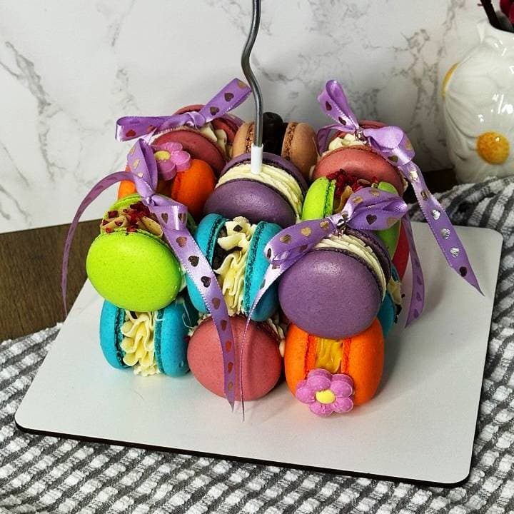 A Macarons Cake Cube 18pc is arranged on a square plate with purple ribbon bows, featuring colorful macarons filled with creamy delights and some adorned with tiny edible flowers. The display sits on a checkered cloth against a marble background.