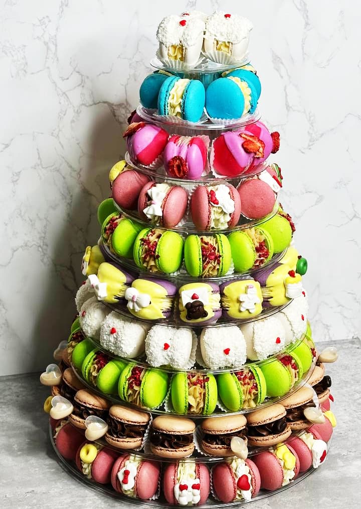 The 10-tier Macaron Tower displays a pyramid of colorful macarons and small desserts in vibrant pink, green, brown, and blue hues, enhanced by white cream-filled treats and decorative flowers.