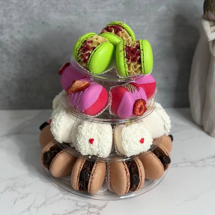 A three-tiered macaron display showcases an array of desserts. The top tier features green macarons with a filling, the middle tier holds pink and red frosted round cakes, and the bottom tier presents brown macarons alongside white coconut-covered treats.