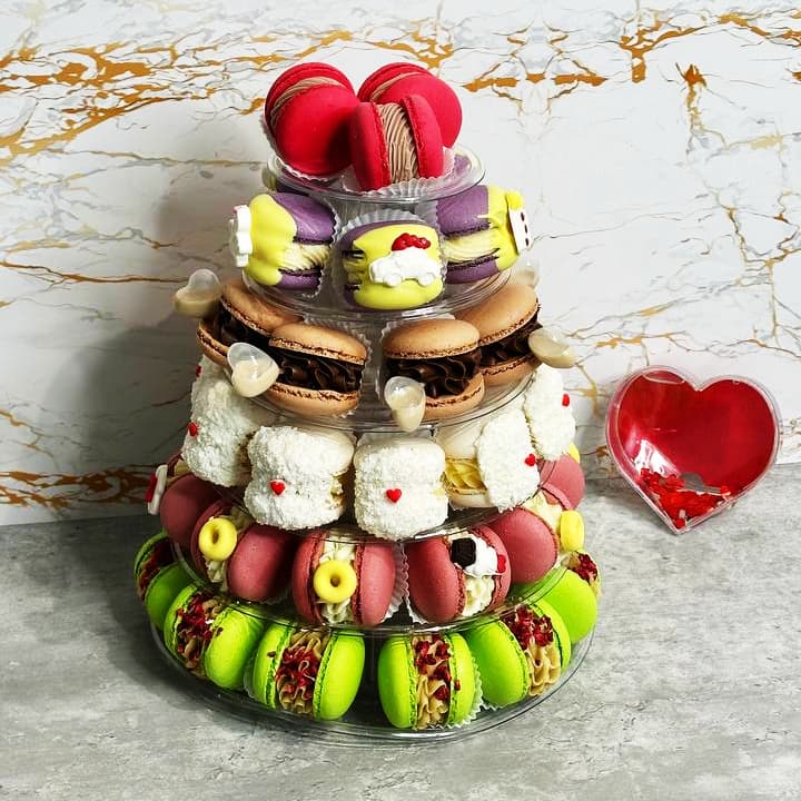 A 6-tier Macaron Tower brimming with colorful macarons, chocolates, and pastries is a sight to behold. To the right sits a small red heart-shaped glass dish filled with red beads, set against a chic marble-patterned wall.