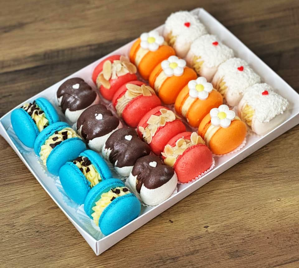 A custom white box containing four rows of colorful macarons rests on a wooden surface. These delightful macarons are adorned with various toppings, including almonds, chocolate drizzles, and floral designs, creating a feast for the eyes and taste buds.