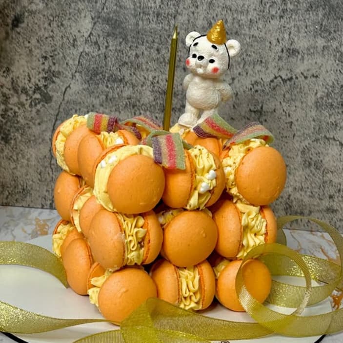 A birthday cake made of stacked orange macarons with yellow filling. There's a small white bear figurine on top wearing a golden party hat. The cake is adorned with a single tall candle and a gold ribbon. The background is a textured gray surface.