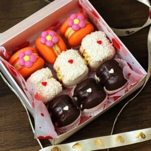 The Сricut Macaron Box showcases nine colorful macarons: orange with pink flowers on top, white with coconut flakes in the middle row, and dark chocolate with small white hearts at the bottom.