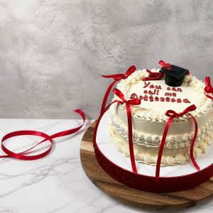 A frosted cake with red ribbons and a graduation cap decoration, with You can call me Doctor written on top, sits on a wooden board.
