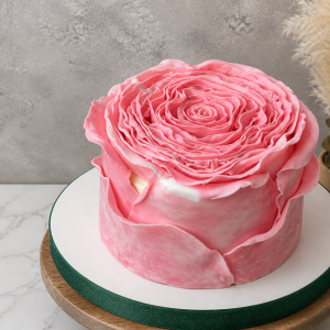 A round cake decorated with pink, petal-like icing to resemble a large flower, placed on a white and green cake board.
