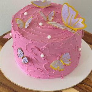 A pink iced cake decorated with white edible pearls and paper butterflies on a round wooden board.