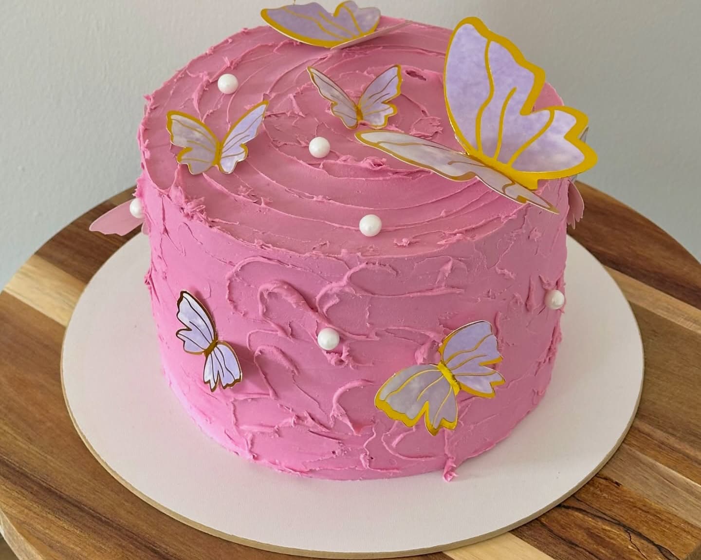 A pink iced cake decorated with white edible pearls and paper butterflies on a round wooden board.