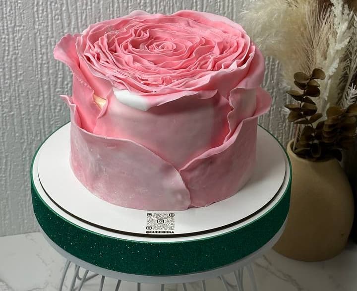 A round cake decorated with pink icing shaped like a large flower, displayed on a green and white cake stand.