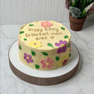 A round cake with yellow icing, colourful flower decorations, and “Happy bday to the best mum ever!” written on top.