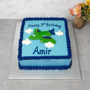 Square blue cake with an aeroplane, clouds, and Happy 3rd Birthday Amir written on top. Vase with orange flowers is beside the cake.