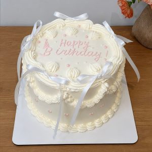 A round white iced birthday cake with pink writing, white ribbons, and decorative piping sits on a white board.