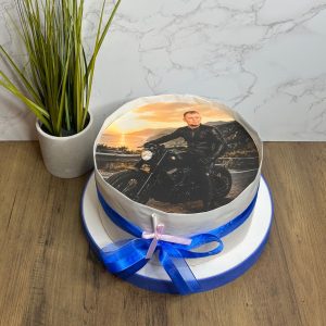 A round cake with a printed photo of a man on a motorbike, decorated with blue and purple ribbons, sits on a wooden surface.