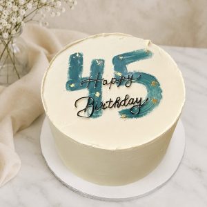 A round cake with 45 and Happy Birthday written on white icing, next to a vase of small white flowers.