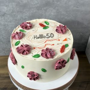 Round cake with white icing, pink and red flower decorations, green leaves, and Hello 50 written on top, on a white stand.
