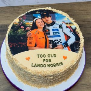 A cake with an image of two people and the text TOO OLD FOR LANDO NORRIS on an orange plaque, with two small orange hearts.