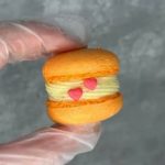 A gloved hand holds an orange macaron filled with cream and topped with two small pink heart decorations on the cream.