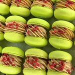 Green macaroons with light brown filling and small red pieces arranged in neat rows on a flat surface.