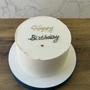 A round white cake with Happy Birthday written on top and a small red heart decoration, placed on a wooden table.