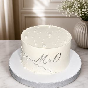 A round white cake with pearl-like decorations, M & O written on the side, sits on a white board near a vase of white flowers.