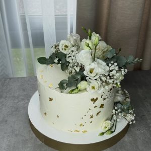 A white cake with gold leaf accents, decorated with white flowers and greenery, sits on a round board beside a window with sheer curtains.