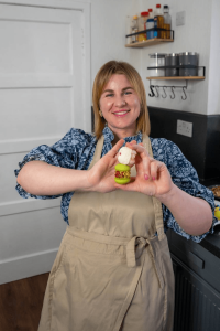 A woman in an apron stands in a kitchen, smiling and holding up a small, decorated pudding with both hands.
