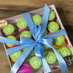 A box of cupcakes with green and white tree logos on top, tied with a blue ribbon, sits on a wooden surface.