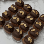 Eighteen chocolate-covered treats topped with peanuts are arranged in rows on a white marble surface.