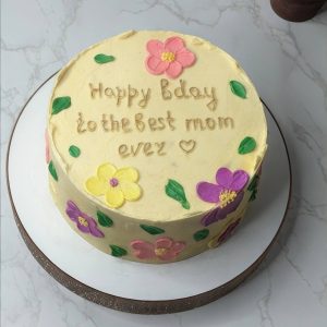 Round cake with pastel flowers and the message Happy bday to the best mum ever written in brown icing on top.