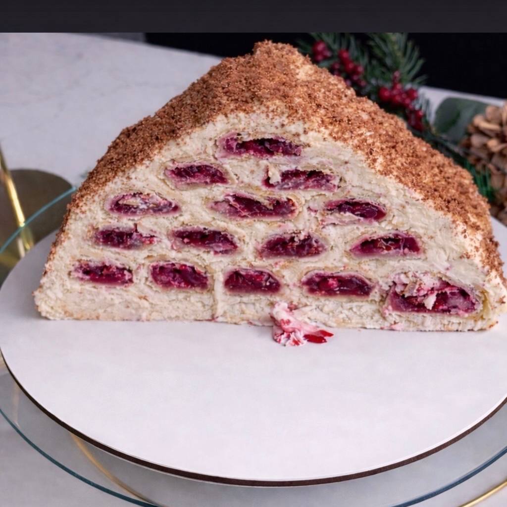 A triangular slice of layered cake with visible cherry filling and crumble topping, displayed on a glass plate with festive decor in the background.