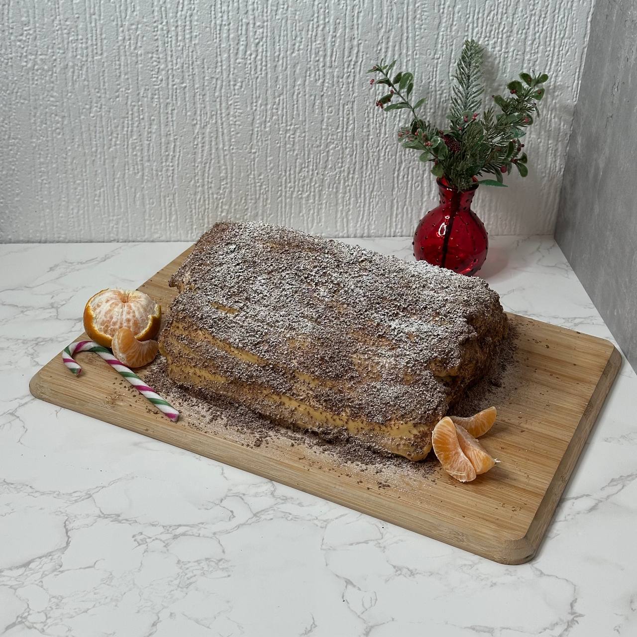 A rectangular layered cake on a wooden board, dusted with cocoa and icing sugar, with tangerine segments and candy canes nearby.