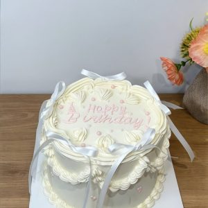 A white iced birthday cake with piped decorations, white ribbons, and Happy Birthday! written in pink, on a wooden table.