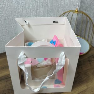 A white gift bag with white handles sits on a wooden floor near a gold wire birdcage and a textured white wall.