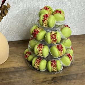 A tiered stand showcases green macarons filled with tan cream and topped with red crumbles, reminiscent of a Scottish treat. Nestled on a wooden surface against a textured white wall, with part of a vase visible on the left, it's the perfect display to inspire you to buy or gift in an elegant box.