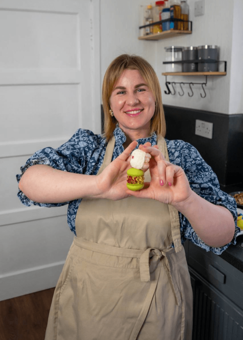 A person wearing a beige apron and a blue patterned shirt holds up a small, decorated cupcake in a kitchen filled with macarons. Shelves with jars and a door are visible in the background, hinting at their bustling UK delivery service.