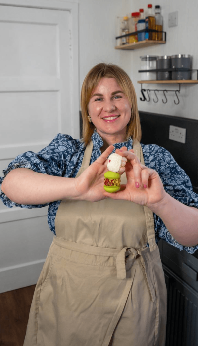 A woman in an apron stands in a kitchen, smiling and holding up a small, decorated pudding with both hands.