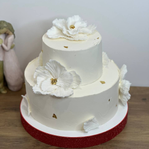 Two-tier white cake with white flower decorations, gold accents, on a red glitter base, displayed on a wooden surface.