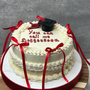 A white iced cake with red ribbons and a small black graduation cap; text on cake reads, You can call me Doctorate.