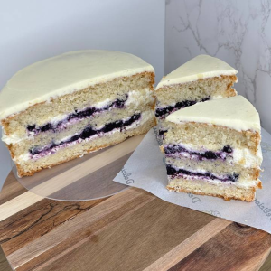 A sliced vanilla sponge with white icing and purple berry filling on a round wooden board, next to a piece of greaseproof paper.